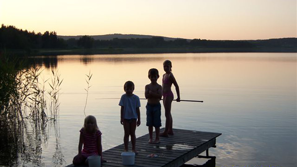 Att fiska är både roligt och spännande, även för de yngre. På bryggan sitter och står tre barn och tittar på sin kompis som metar. Sjön Summeln ligger i västra delen av Kila socken i Säffle kommun.