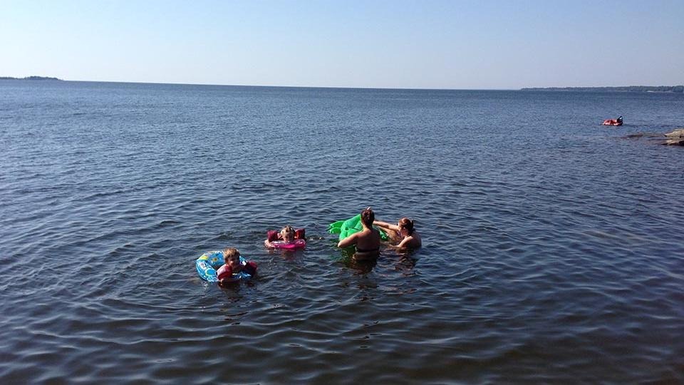 Här på bild ses både unga och vuxna som badar och leker i Vänern, de har badmadrass med sig och färgglade flytringar på sig.
