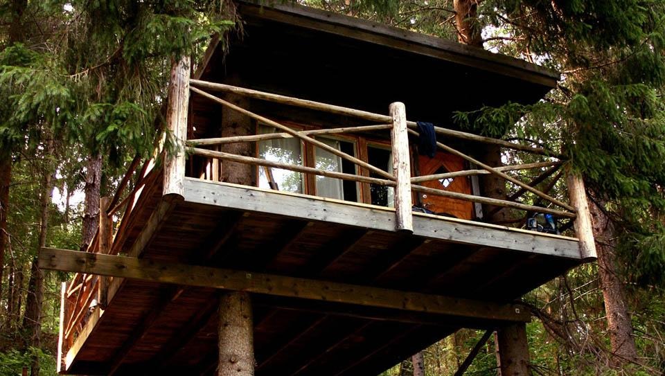 Foto på en timrad stuga som sitter högt upp bland träden med altan för utsikt över sjön Eldan. Denna kallas Gökboet här kan du sova gott till suset bland trädtopparna. 
