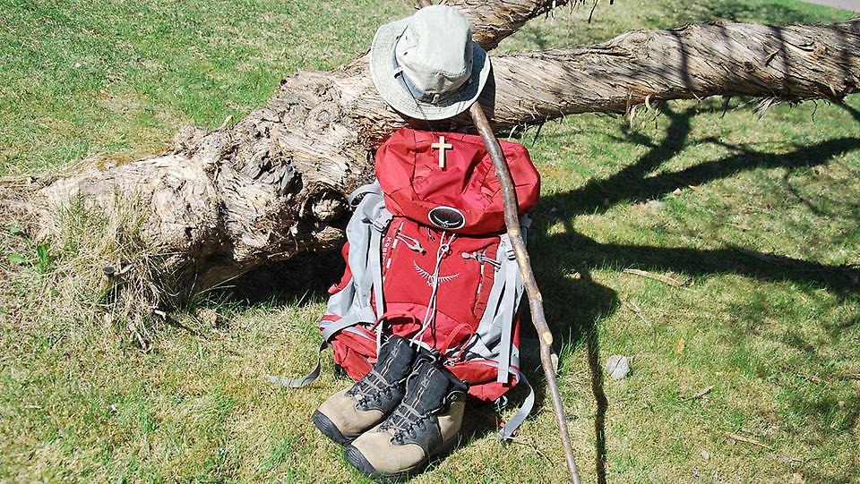 På bilden är det vandringskängor, hatt, vandringsstav och en röd ryggsäck där hänger ett träkors i ett band.
